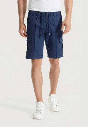 Uomo che indossa pantaloncini cargo blu navy con coulisse e bottone, scarpe da ginnastica bianche, in piedi su sfondo uniforme.