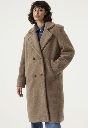 Vrouw die een lange bruine teddyjas met grote revers draagt over een blauw denimshirt en spijkerbroek, poseert met één hand in de jaszak.