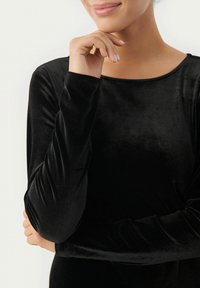Haut en velours noir à manches longues avec un col rond. Le tissu a une texture lisse et un léger éclat, accentué par des manches ajustées.