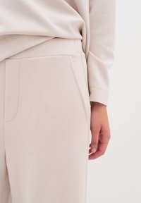 Pantalons de survêtement beige clair avec une texture lisse, des poches latérales et une taille ajustée, avec un haut beige clair partiellement visible.