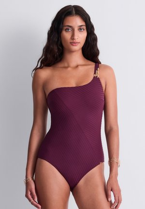 Femme aux longs cheveux foncés portant un maillot de bain violet texturé asymétrique avec des accents dorés et de fines bracelets dorés aux deux poignets.