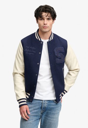 Ung mand med mørkt krøllet hår iført en navy- og cremefarvet varsityjakke over en hvid skjorte og blå jeans, stående med den ene hånd i lommen.