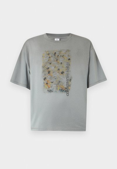 Γκρι μεγάλης διάστασης T-shirt με ξεθωριασμένο αφηρημένο σχέδιο πλήθους και κάθετο κείμενο "WON HUNDRED" στο κέντρο μπροστά.