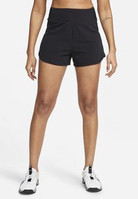 Mujer vestida con pantalones cortos deportivos negros y zapatillas de correr blancas, de pie con las piernas abiertas sobre un fondo blanco liso.