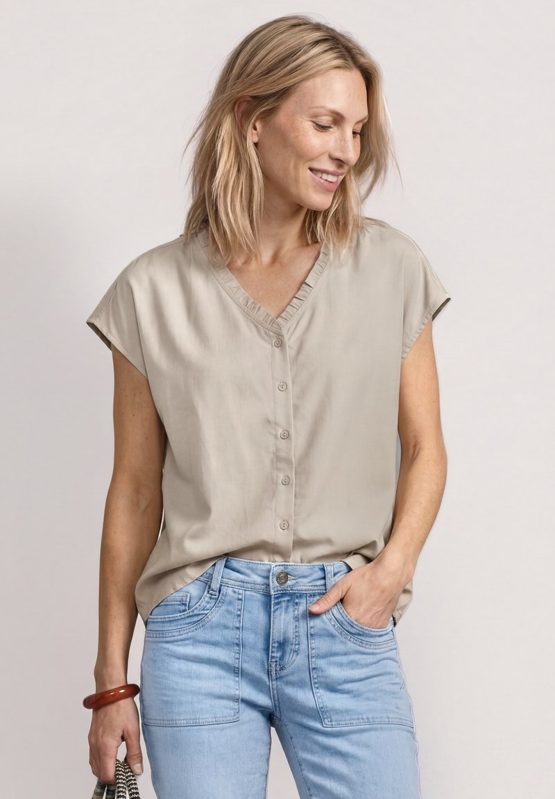 Femme aux cheveux blonds portant un chemisier beige à manches courtes boutonné et un jean bleu clair, souriante et regardant vers le bas, une main dans la poche.