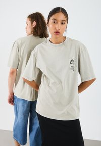 Helles graues, übergroßes T-Shirt aus Baumwolle, kurze Ärmel, mit bedruckten Charakteren auf der Vorderseite. Getragen von zwei Personen, von denen eine von hinten zu sehen ist.