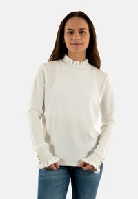 Pullover bianco con colletto alto arricciato, dotato di motivi a pallini e polsini a balze, accentuati da tre bottoni su ciascun manica.