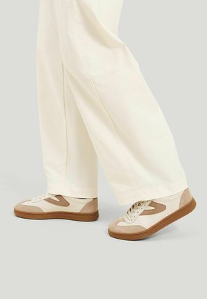 Person iført cremefarvede, lige bukser og beige og brune lave sneakers træder fremad.