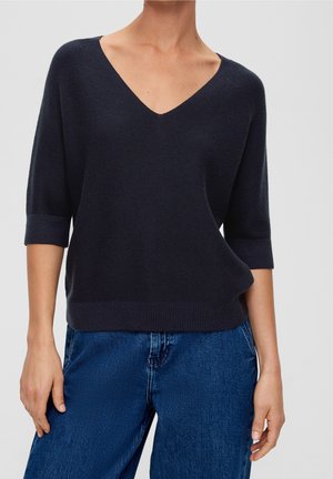 Femme portant un pull en maille à col en V bleu marine foncé avec des manches jusqu'au coude et un jean en denim bleu sur un fond uni.