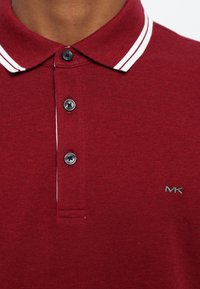 Bordeaux polo skjorte med hvit-stripet krage, med tre svarte knapper og et brodert "MK"-logo nederst til høyre.