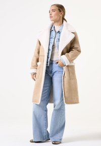 Manteau oversize beige et crème avec une texture pelucheuse, grand col et deux poches avant, superposé sur une veste en denim clair et un jean évasé.