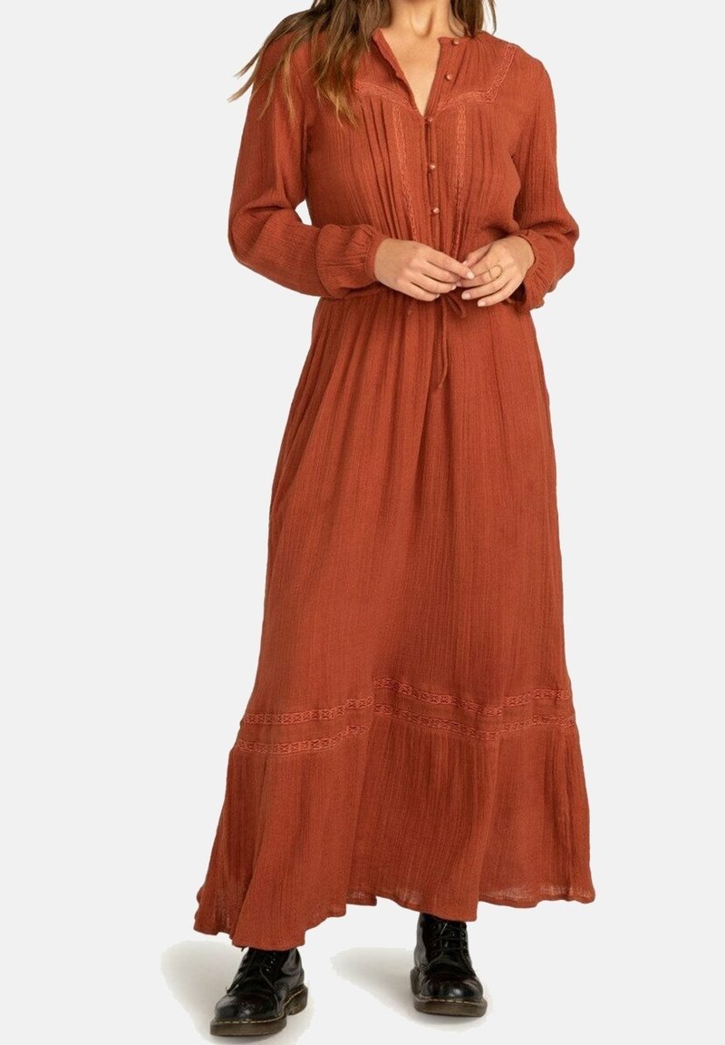 Femme portant une robe longue à manches longues de couleur rouille avec des boutons et des détails en dentelle, associée à des bottes noires à lacets.