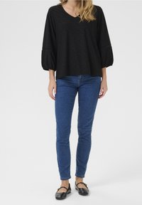 Blouse noire texturée avec un col en V et des manches bouffantes, associée à un jean bleu ajusté et des chaussures plates noires.