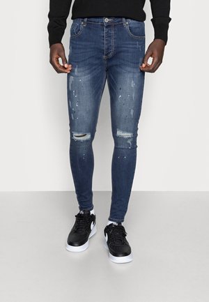 Homme portant un jean slim bleu déchiré avec des éclaboussures de peinture, assorti à des baskets noires et blanches et un haut noir à manches longues.