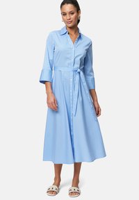 Blau-weiß gestreiftes Hemdkleid, knielang, mit kurzen Ärmeln und durchgehender Knopfleiste, mit gebundenem Taillengürtel, kombiniert mit weißen Sandalen.