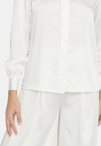 Blusa bianca con un sottile motivo a pois, maniche lunghe e pattina con bottoni, abbinata a pantaloni a gamba larga coordinati. Texture liscia in tutto il capo.