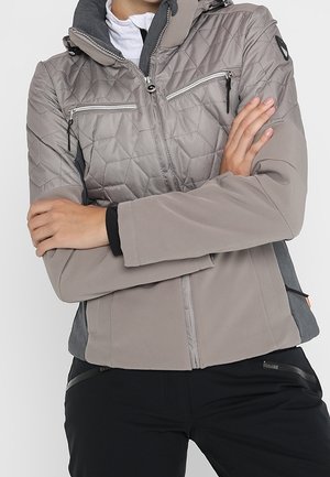 Osoba v světle šedé prošívané bundě s kapsami na zip na hrudi, stojící se založenýma rukama, černé kalhoty viditelné od pasu dolů.