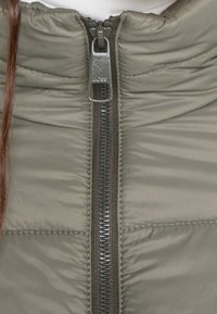 Närbild på dragkedjan på en grå vadderad jacka med "BOSS"-logotyp på den metalliska dragfliken.