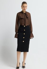 Braune transparente Bluse mit einer Schleife am Hals, kombiniert mit einem schwarzen Bleistiftrock, der goldene Knopfakzente und Seitentaschen aufweist. Schwarze High Heels.