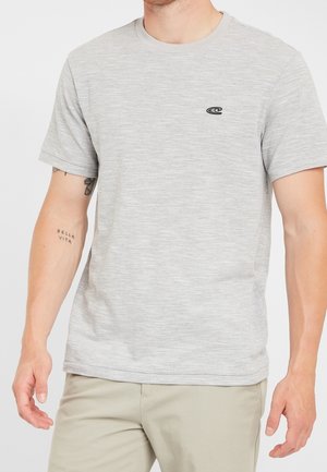 Mężczyzna w jasnoszarej koszulce z krótkim rękawem i małym czarnym logo na piersi oraz beżowych spodniach, pokazujący tatuaż z napisem "Bella Vita" na lewym przedramieniu.