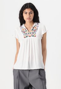 Blouse blanche avec un col en V et des manches courtes à volants. Présente une broderie florale colorée le long de l'encolure et une texture plissée.