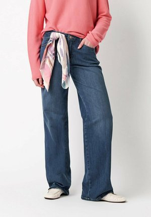 Person wearing wide-leg blue jeans with a pastel patterned scarf belt, coral long-sleeve top, and white slip-on shoes, standing against white background.