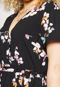 Schwarzes Blumenkleid mit tiefem V-Ausschnitt, kurzen Ärmeln und Taillengürtel. Die Blumen sind in Mustern aus Pink, Weiß, Orange und Türkis.