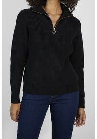 Schwarzer, gerippter Pullover mit halbem Reißverschluss und Stehkragen, ausgestattet mit einem strukturierten Strickmuster und einem goldfarbenen Reißverschlussanhänger. Kombiniert mit blauen Jeans.