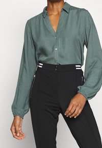 Blouse verte texturée avec col et fermeture à boutons, associée à un pantalon noir taille haute avec des accents rayés argentés et des détails de fermeture éclair.