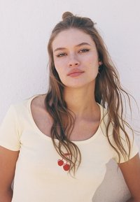Gul bomullst-shirt med rund halsringning och rött körsbärsgrafik på framsidan. Modellen har långt brunt hår och står mot en vit bakgrund.