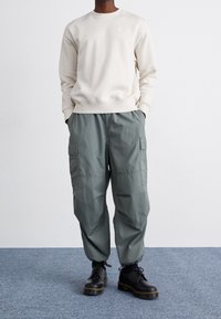 Beige cotton sweatshirt with white Nike logo, paired with green cargo pants featuring side pockets, and black lace-up boots with chunky soles.