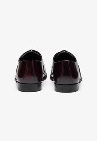 Chaussures en cuir bordeaux brillantes avec une texture lisse, bout arrondi et semelle noire contrastante. Présente un subtil détail de couture et aucun élément en métal.