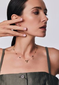 Złoty naszyjnik z czarnymi koralikami i sercowym wisiorkiem. Modelka w oliwkowej bluzce z guzikiem, trzymająca ucho przy twarzy.