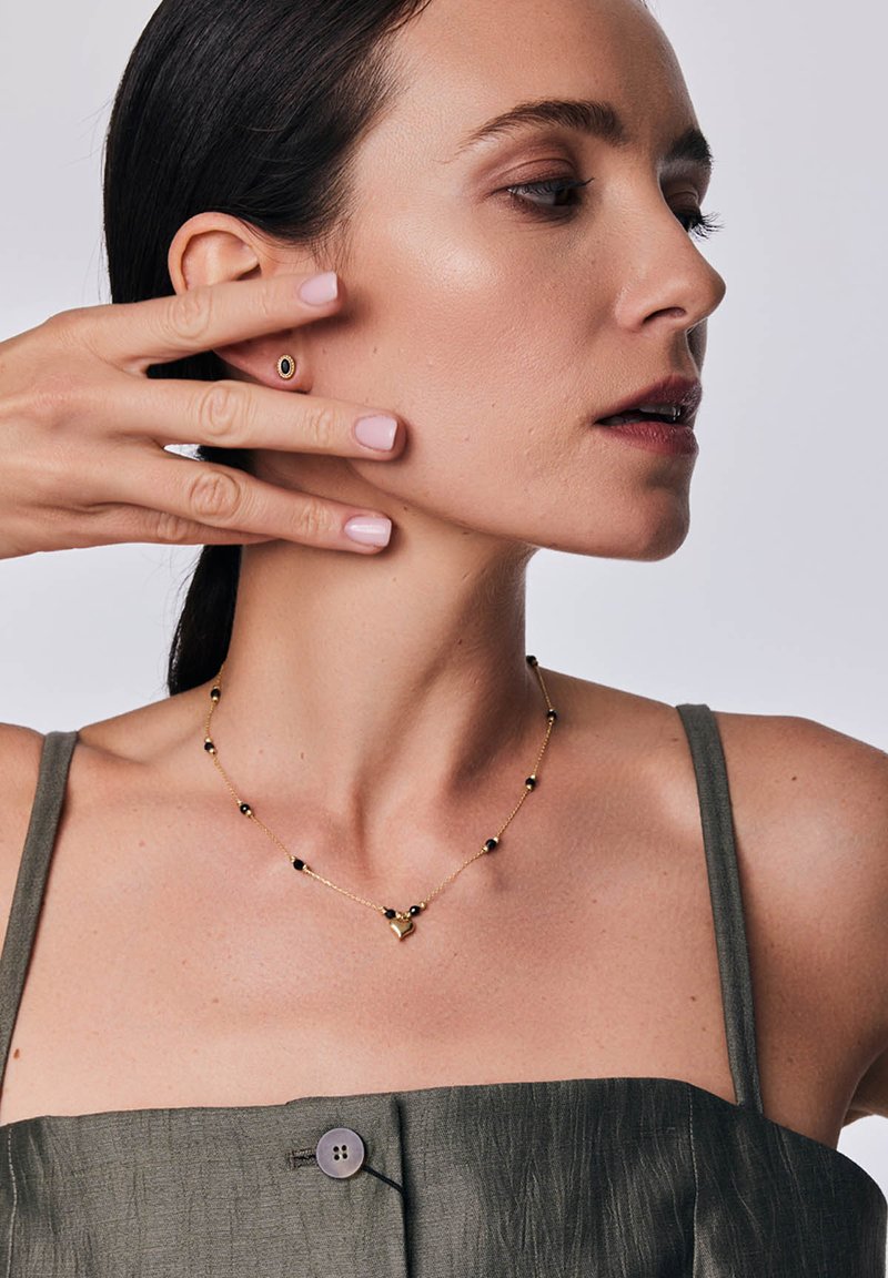 Złoty naszyjnik z czarnymi koralikami i sercowym wisiorkiem. Modelka w oliwkowej bluzce z guzikiem, trzymająca ucho przy twarzy.