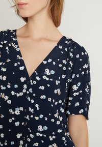 Abito floreale blu navy con scollatura a V, maniche corte e chiusura a bottoni. Il tessuto ha una trama morbida con piccoli fiori bianchi, blu e beige.