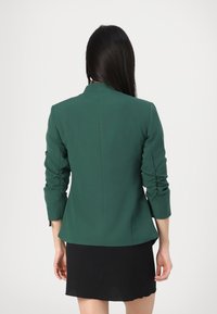 Tmavě zelený šitý blazer s nařasenými tříčtvrtečními rukávy, přiléhavým střihem a čistými liniemi. Nosený přes černou sukni.