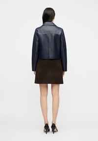 Femme portant une veste en cuir courte bleu marine, une jupe marron foncé et des chaussures à talons noirs, debout de dos sur un fond uni.