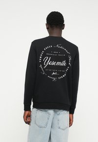 Felpa nera con testo bianco sul retro, che include "Yosemite," circondato da elementi grafici circolari e orlo a costine.