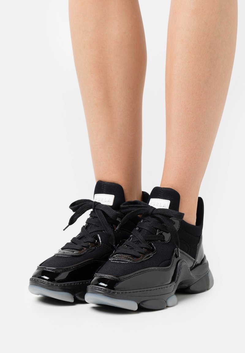 Jambes portant des baskets montantes noires avec des détails en cuir verni, des semelles épaisses et des lacets noirs sur un fond blanc.