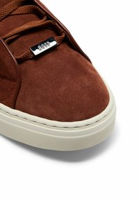 Brown suede sneaker featuring a white rubber sole, textured detailing, and silver hardware on the laces. Simple and modern design.