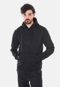 Felpa nera realizzata in tessuto morbido, con tasca a marsupio e cappuccio regolabile. Presenta maniche lunghe e una vestibilità comoda. Indossata con jeans scuri.