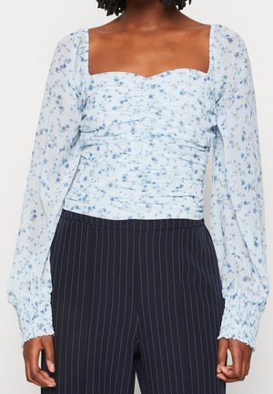 Vrouw die een lichtblauw gebloemd gerimpeld blouse met lange mouwen en een vierkante hals draagt, met marineblauwe pantalon met fijne streepjes.