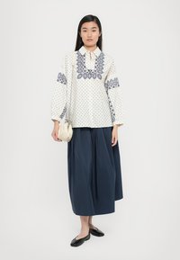Weiße Bluse mit navyblauen Punkten und floraler Stickerei, kombiniert mit einem langen, navyblauen Rock. Als Accessoires dienen eine cremefarbene, weiche Handtasche und schwarze Ballerinas.