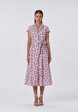 Femme portant une robe midi fleurie rose à manches courtes, col, boutonnée sur le devant, ceinture à la taille et talons blancs à brides, debout devant un fond uni.