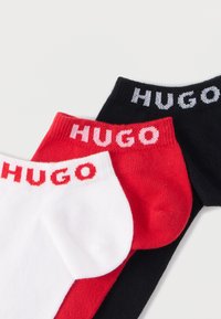 Kolme paria nilkkasukkia väreissä valkoinen, punainen ja musta, joiden resorissa on "HUGO"-teksti vastaavilla väreillä. Valmistettu pehmeästä kankaasta.