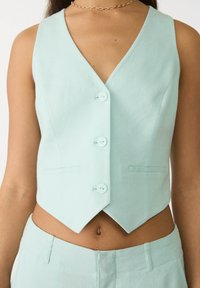 Miętowa kamizelka z dekoltem w kształcie litery V, pięcioma guzikami i dwoma przednimi kieszeniami. Materiał ma gładką fakturę i dopasowany krój.