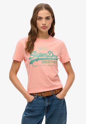 T-shirt en coton rose avec le texte imprimé vert "REAL SuperDry" et un accent décoratif. Manches courtes et encolure ronde. Coupe décontractée.