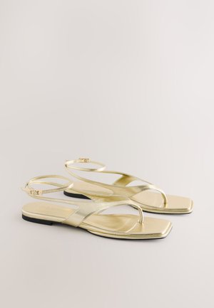 Guldmetallic flade sandaler med et minimalistisk design, tynde ankelremme og firkantet tå. Sålen er sort og kontrasterer den skinnende overdel.
