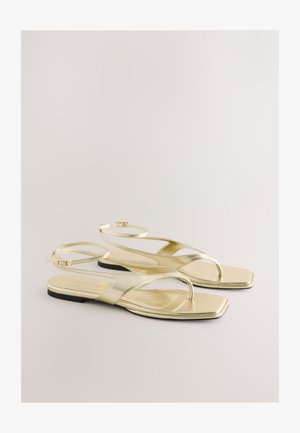 Guldmetallic flade sandaler med et minimalistisk design, tynde ankelremme og firkantet tå. Sålen er sort og kontrasterer den skinnende overdel.
