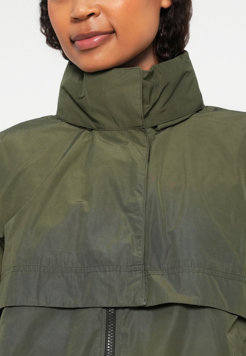 Olivgrüne Jacke mit hohem Kragen, Reißverschluss und wasserabweisendem Material mit glatter Textur. Auffällige Nähte sind sichtbar.